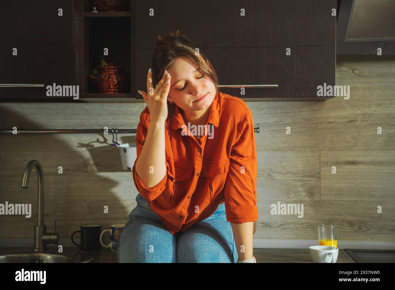 Sad young woman sitting on the kitchen countertop. Portrait of a young housewife looking worried, tired and overworked. Stock Photo