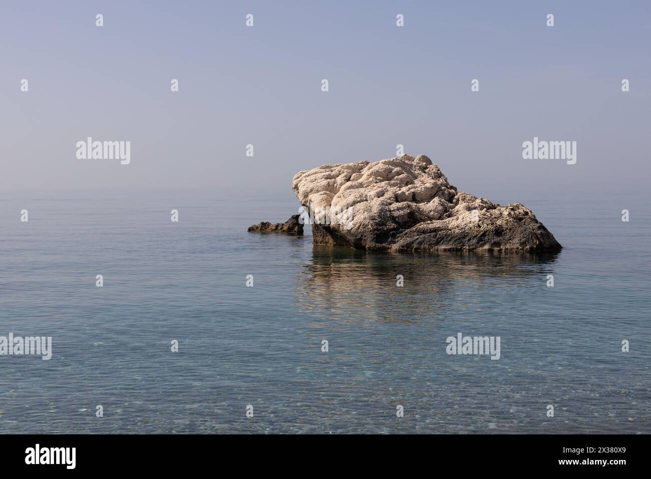 Weathered Rock in Serene Sea. Clear blue waters of the Mediterranean Sea, under a soft sky. Stock Photo