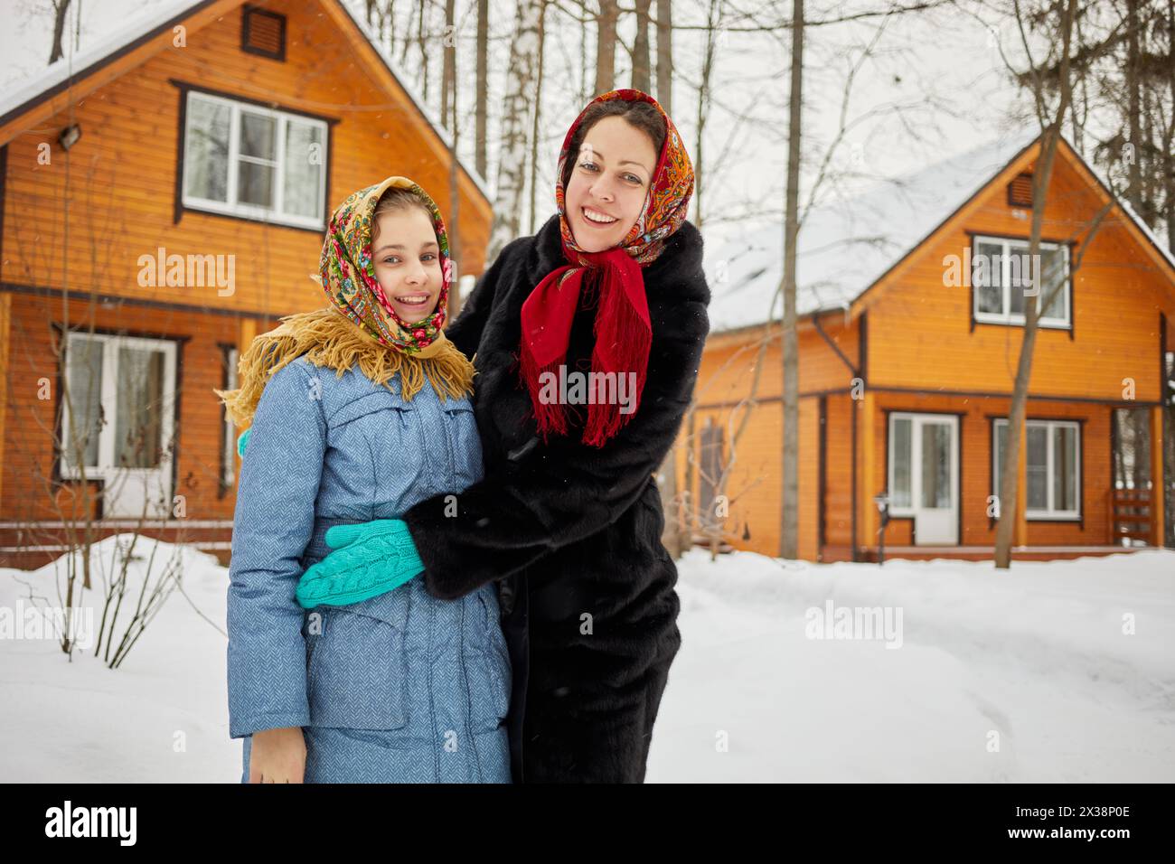 Smiling woman and teenage girl in folk style shawls stand against wooden houses on winter day. Stock Photo