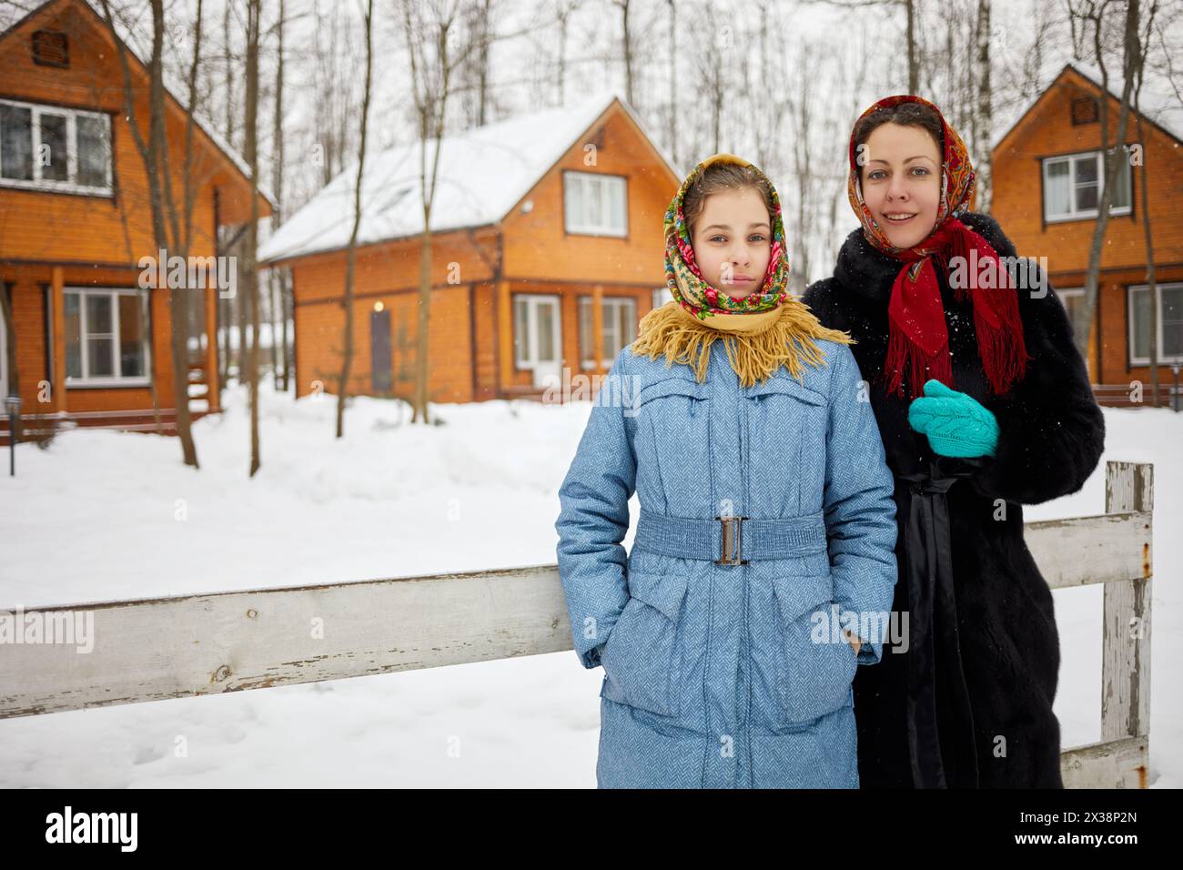 Smiling woman and teenage girl in folk style shawls stand near fencing against wooden houses on winter day. Stock Photo