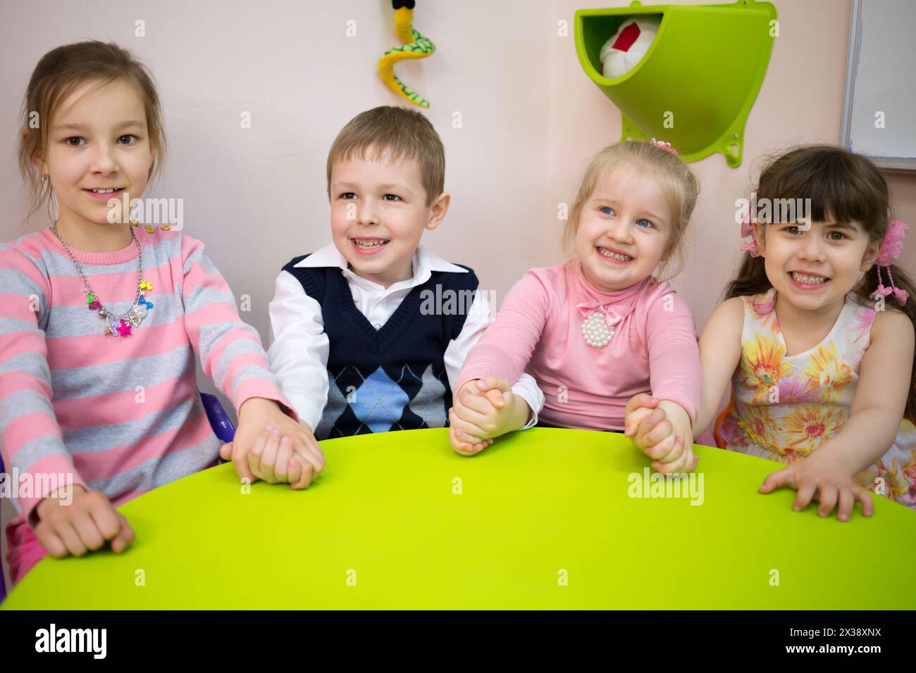 Four little girls and boy are sitting at the table and holding hands. Stock Photo