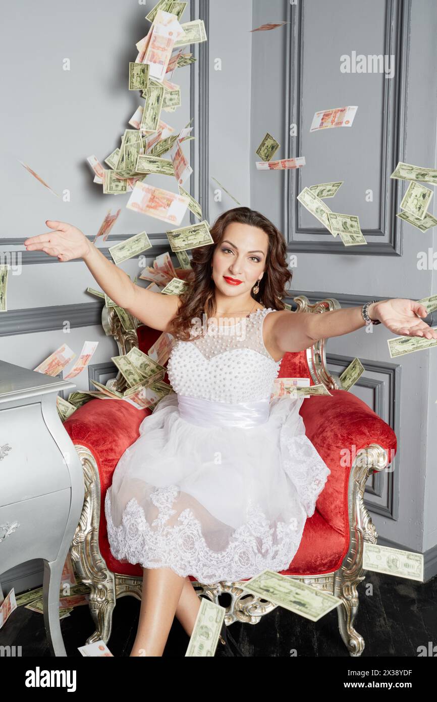 Woman in white dress sits in armchair at room under money banknotes shower. Stock Photo