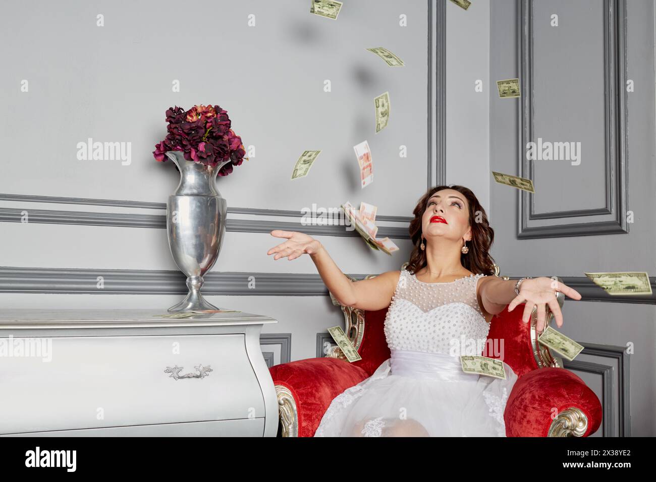 Woman in white dress sits in armchair at room under money banknotes shower. Stock Photo