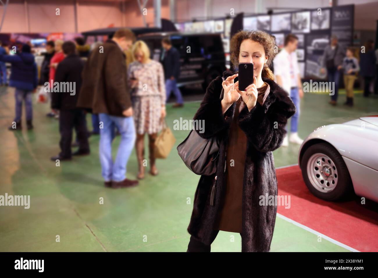 Happy pretty woman in fur coat shoots by phone at exhibition Stock Photo