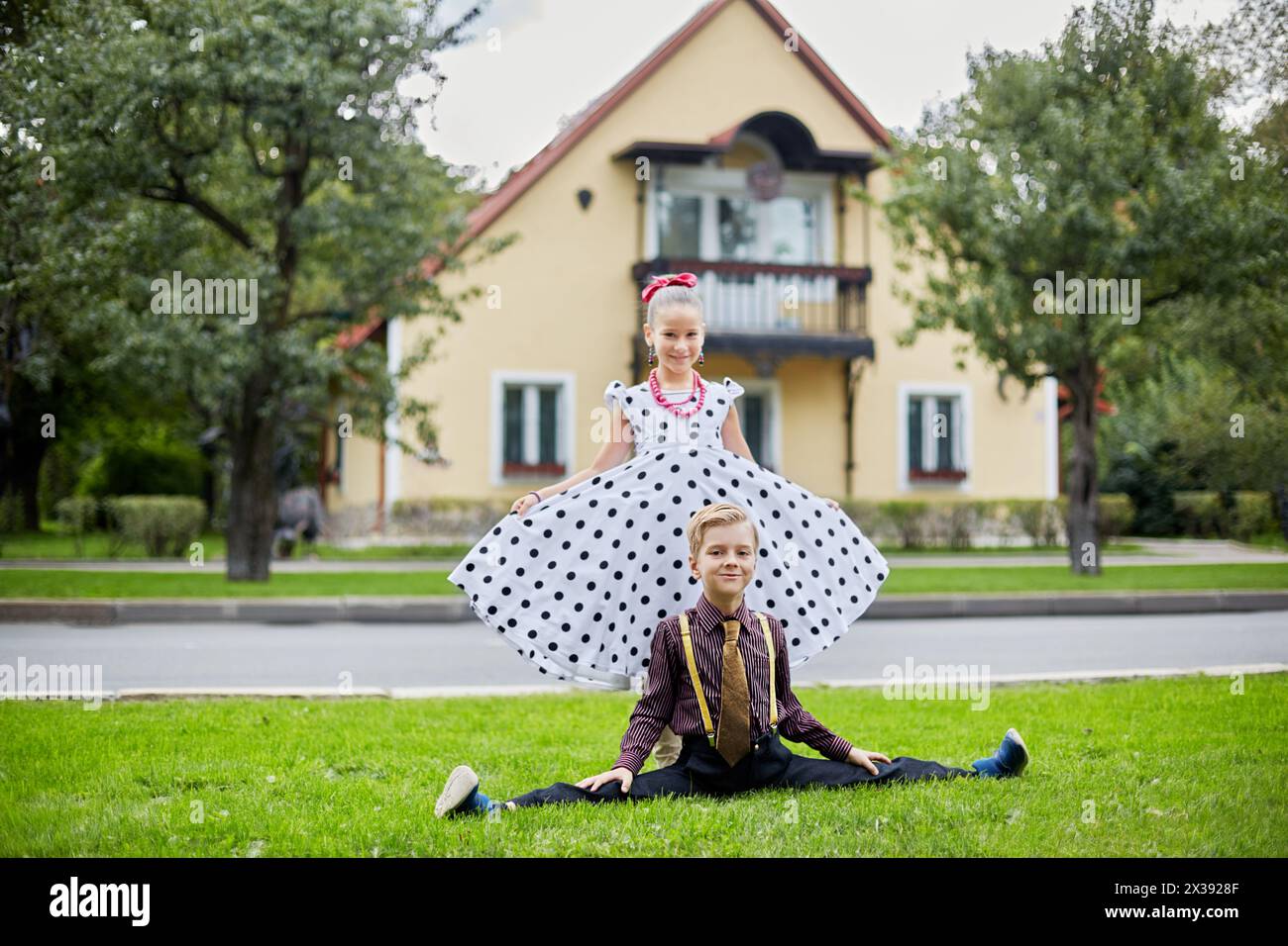 Smiling boy sits on side split and girl in polka-dotted dress stands behind him on grassy lawn against two-storied house. Stock Photo