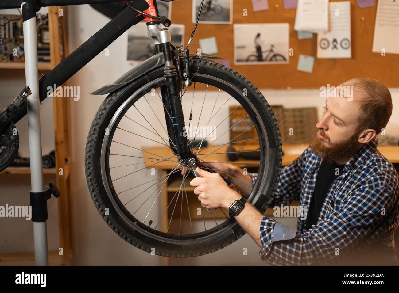 Bearded red-haired man in a checkered shirt repairs a bicycle while squatting in a garage or workshop Stock Photo
