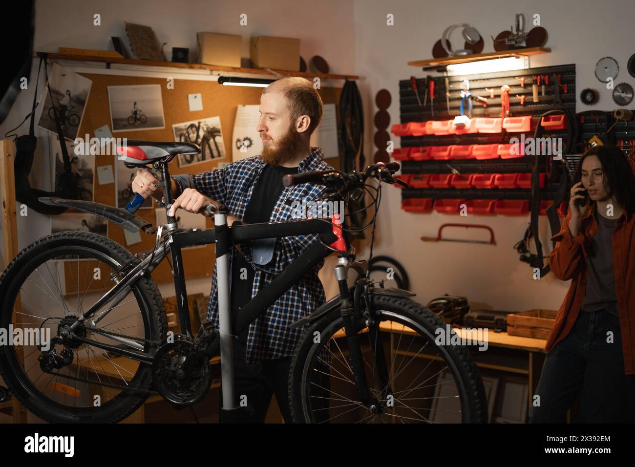 Male mechanic repairing a bicycle in a workshop or garage, his woman waiting in the background. Bike Maintenance Stock Photo
