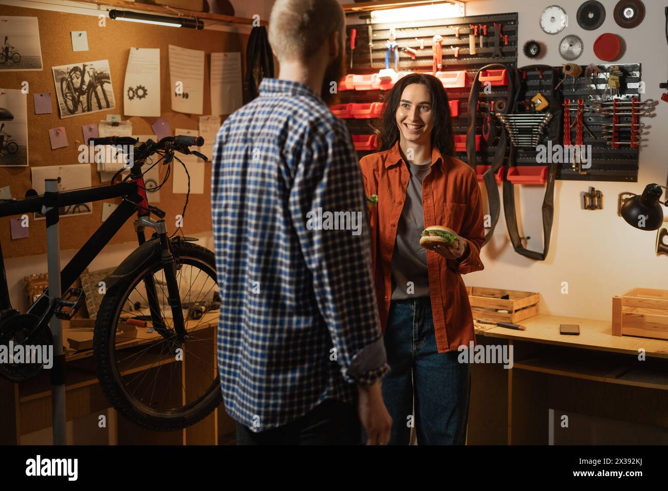 Couple bicycle mechanic in repair workshop. In cycle garage man and woman eating burgers on lunch. Bike Maintenance Stock Photo