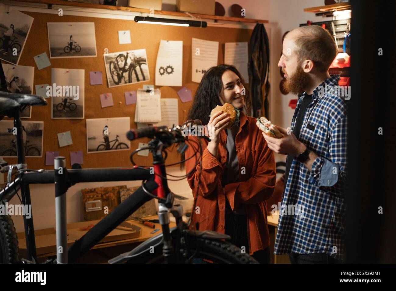 Young couple having a cheerful conversation and eating delicious hamburgers while standing in a bicycle repair shop. Colleagues having lunch in garage Stock Photo