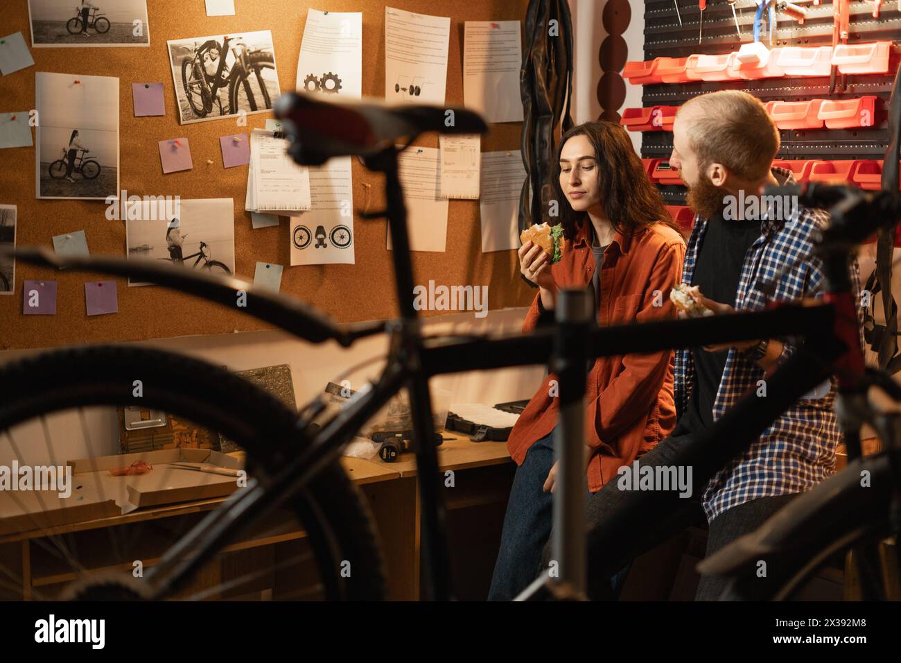 Couple in love chats happily and eats delicious burgers after fixing and servicing a bicycle in repair shop. People working and eating in garage Stock Photo