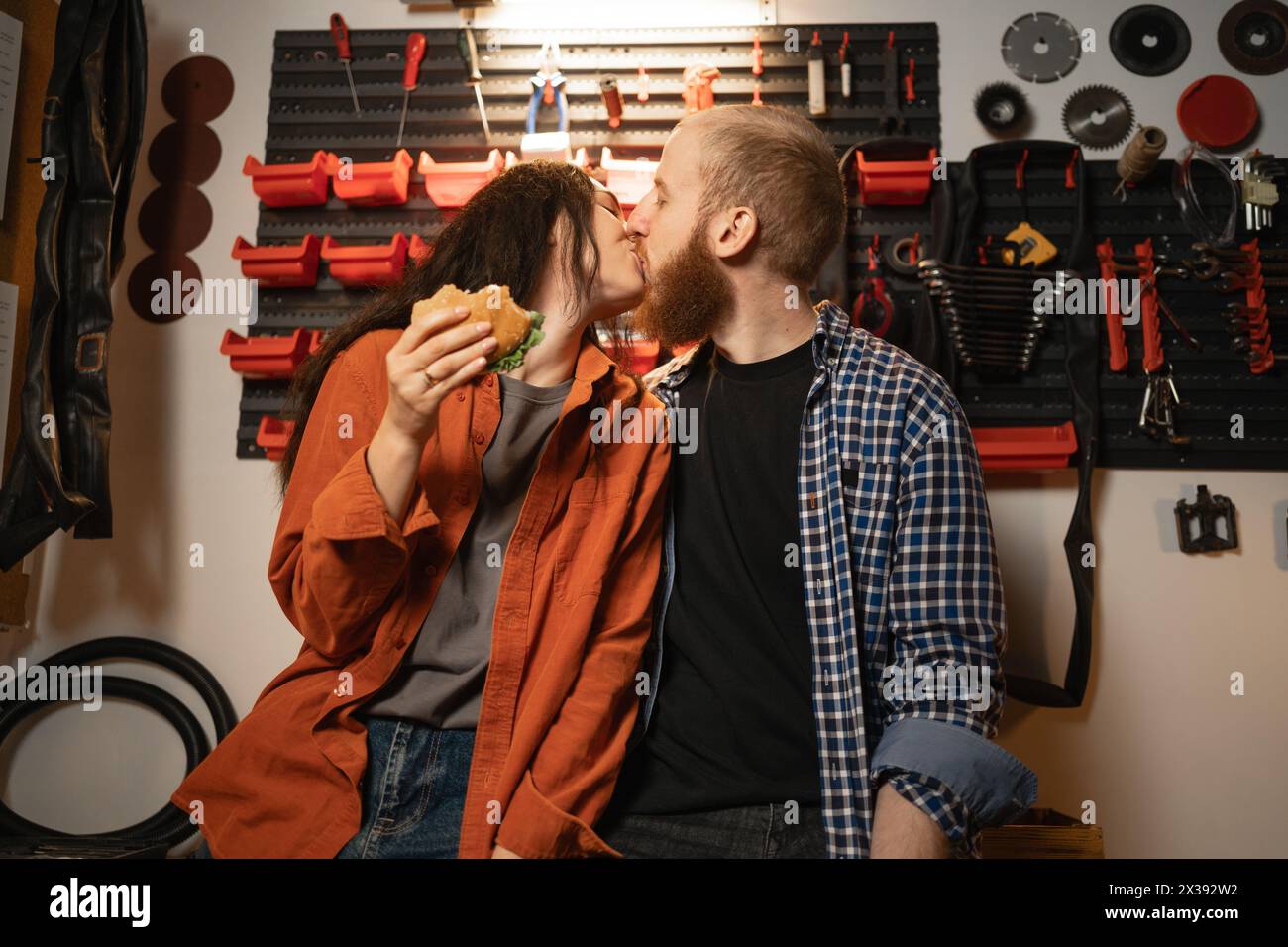 Kissing couple in garage, man and woman eating burgers and kissing after renovation in workshop. Stock Photo
