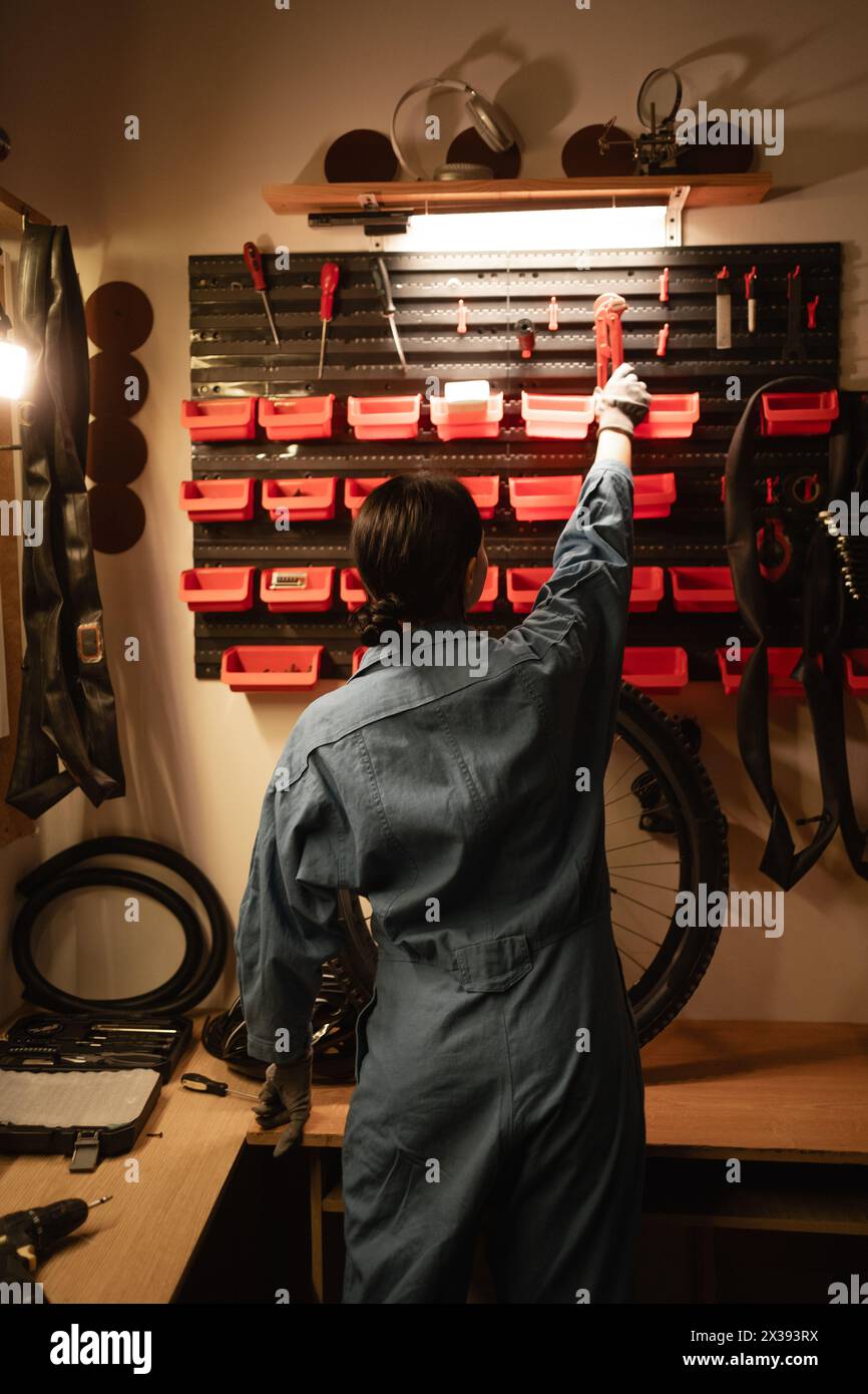 Rear view of a brunette bicycle mechanic working in her garage, standing in front of a wall with tools and equipment for repairs Stock Photo