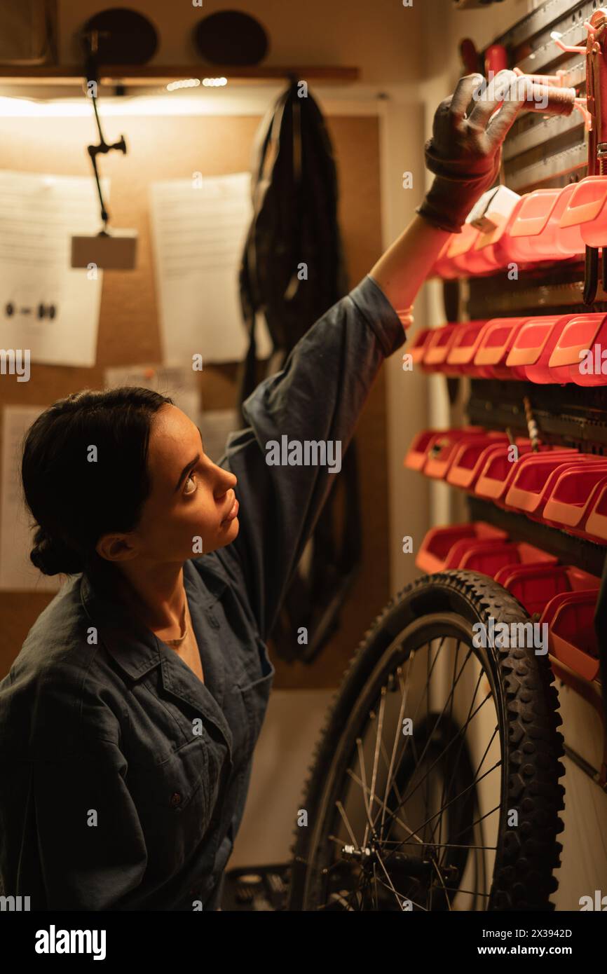 A bicycle mechanic girl standing near the wall reaches for a tool hanging on the wall. Work in an authentic garage Stock Photo