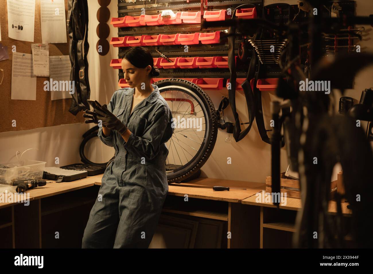 Young professional mechanic woman in blue overalls use mobile phone browsing fix problem in bike repair shop workshop Stock Photo