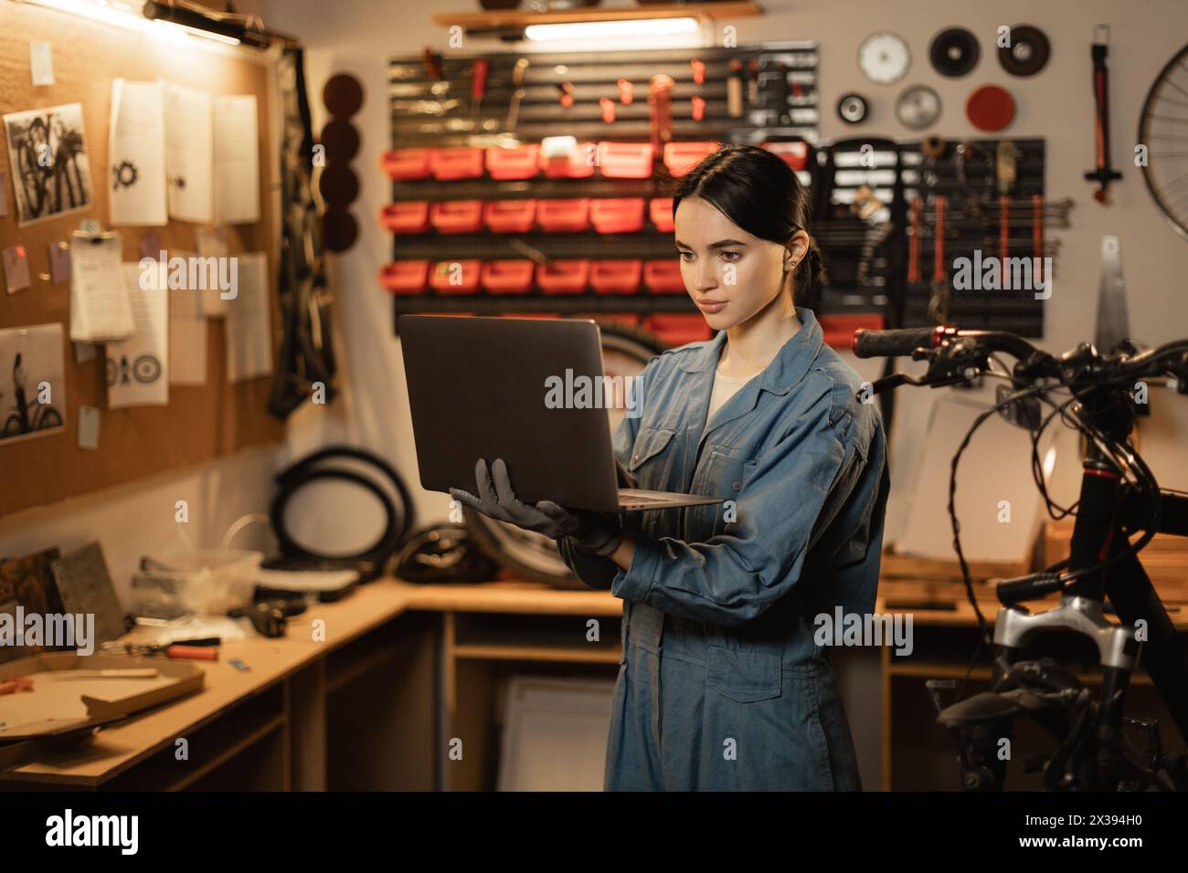 Brunette woman in casual clothes using a laptop and smiling while standing in the bicycle repair shop Stock Photo