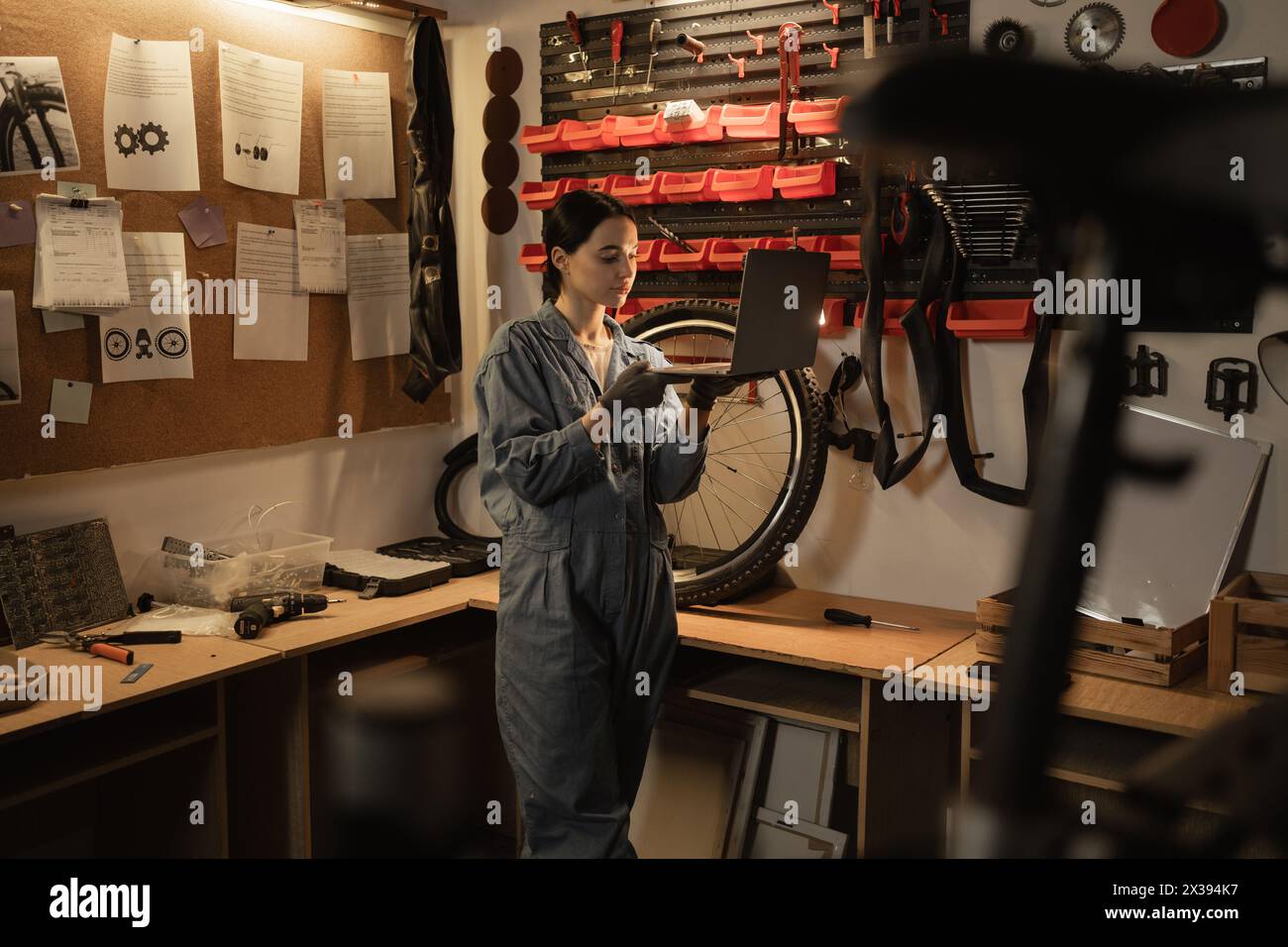 Young female mechanic in bike workshop working and researches information on his laptop. Stock Photo