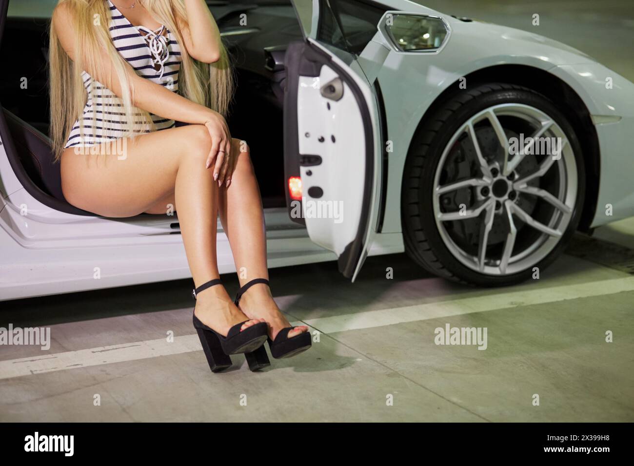 Young blonde woman in striped bodysuit and high-heel shoes poses sitting on passenger seat of modern white car at underground parking. Stock Photo