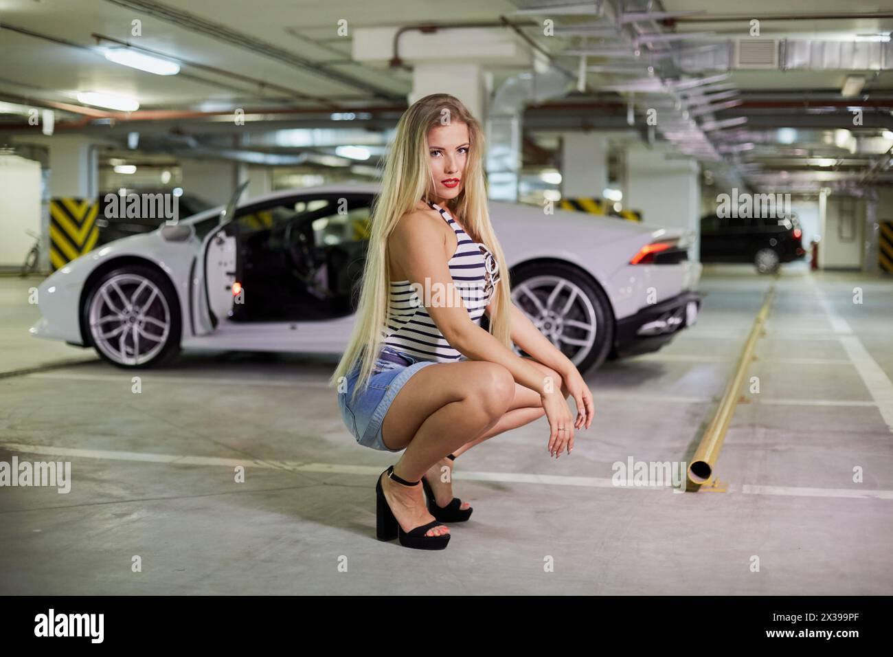 Young smiling blonde woman in striped bodysuit, jeans overall and high-heel shoes poses sitting squatted against modern white car at underground parki Stock Photo