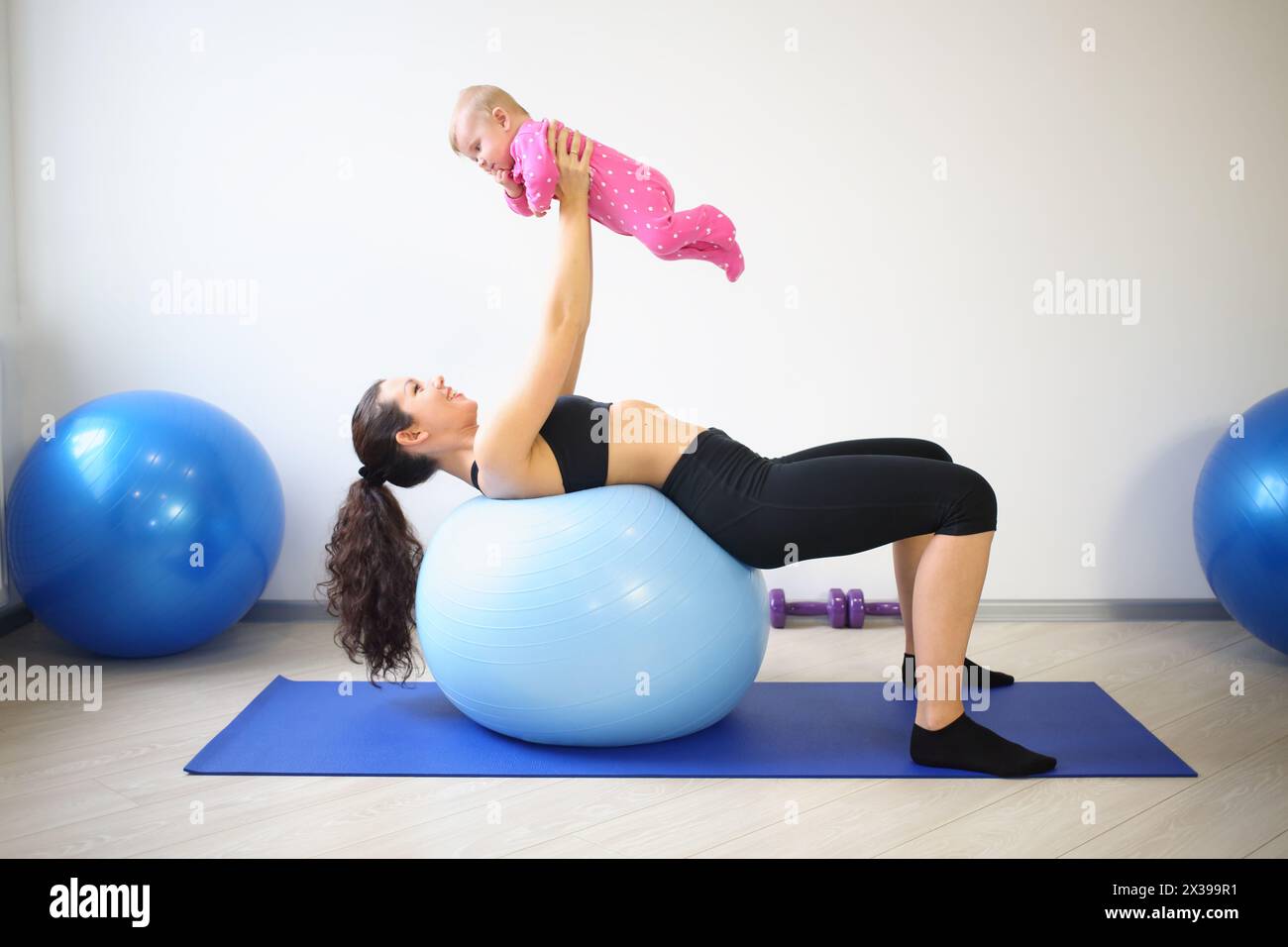 Happy woman lying on the big blue ball on her back lifted up smiling baby in the gym Stock Photo