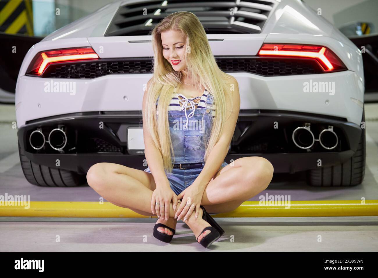 Young woman in striped bodysuit, jeans overall and high-heel shoes sits on yellow pipe at back of modern white car at underground parking. Stock Photo