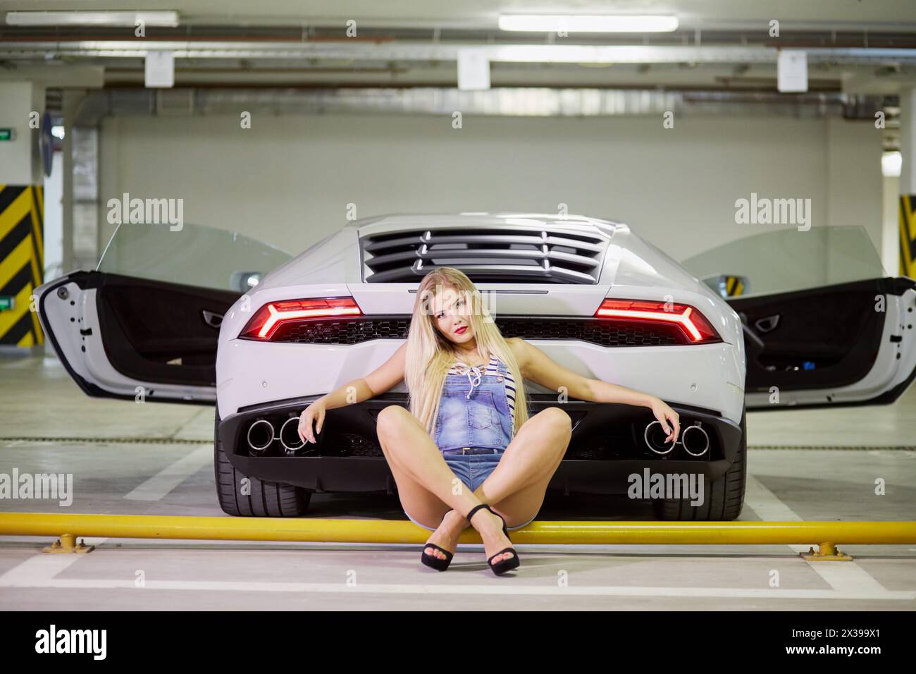 Young woman in striped bodysuit, jeans overall and high-heel shoes sits on yellow pipe leaning at back of modern white car at underground parking. Stock Photo