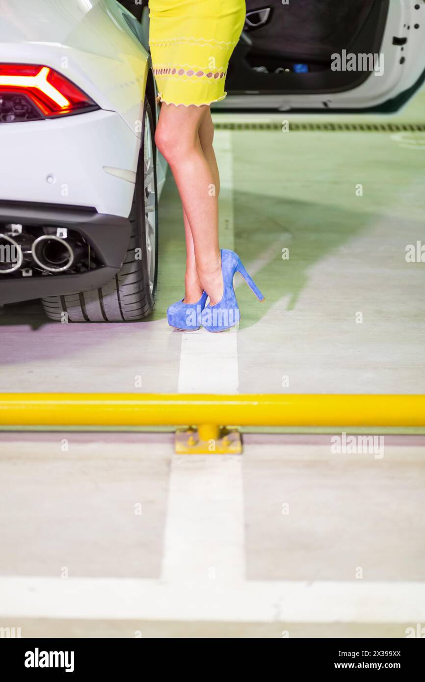 Legs in blue high-heel shoes of woman in yellow dress standing near modern white car. Stock Photo