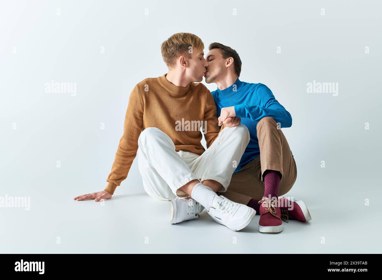Two young men in casual clothes sitting on the ground kissing each other. Stock Photo