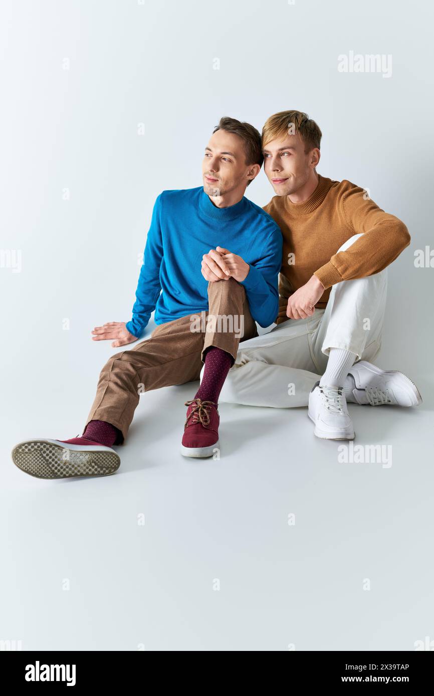 A loving gay couple in casual attires sit closely on a gray backdrop. Stock Photo