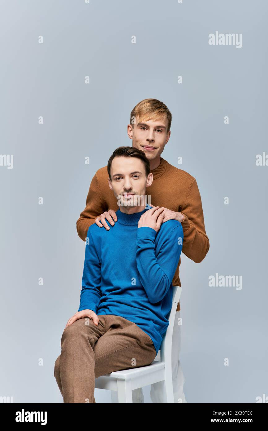 Two loving gay men in casual attire pose for a picture on a white chair against a gray backdrop. Stock Photo