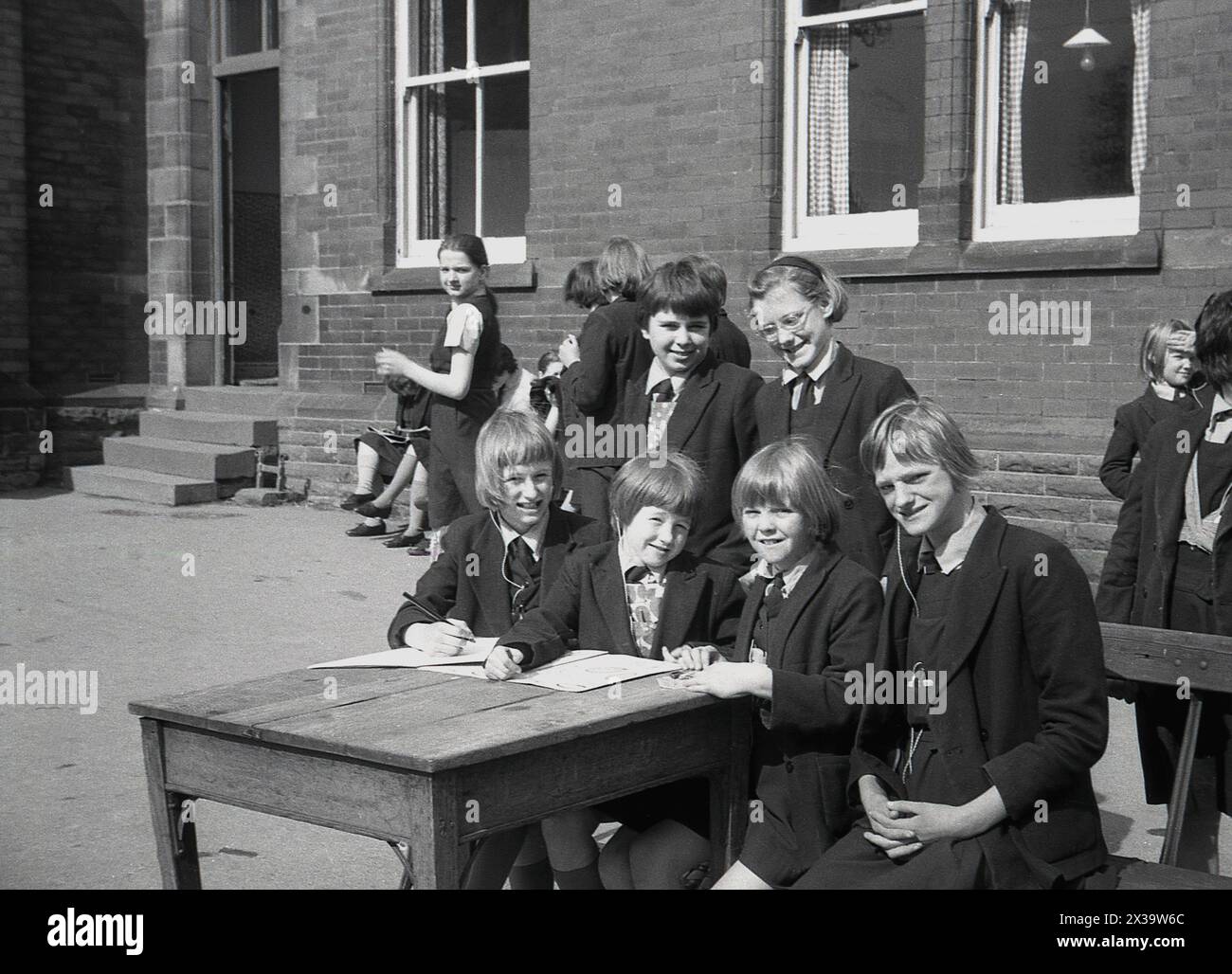 1960s, historical, early summertime and roman catholic schoolgirls in their uniforms sitting outside their school building, with pen and paper, doing their school work at a wooden desk, England, UK. Stock Photo