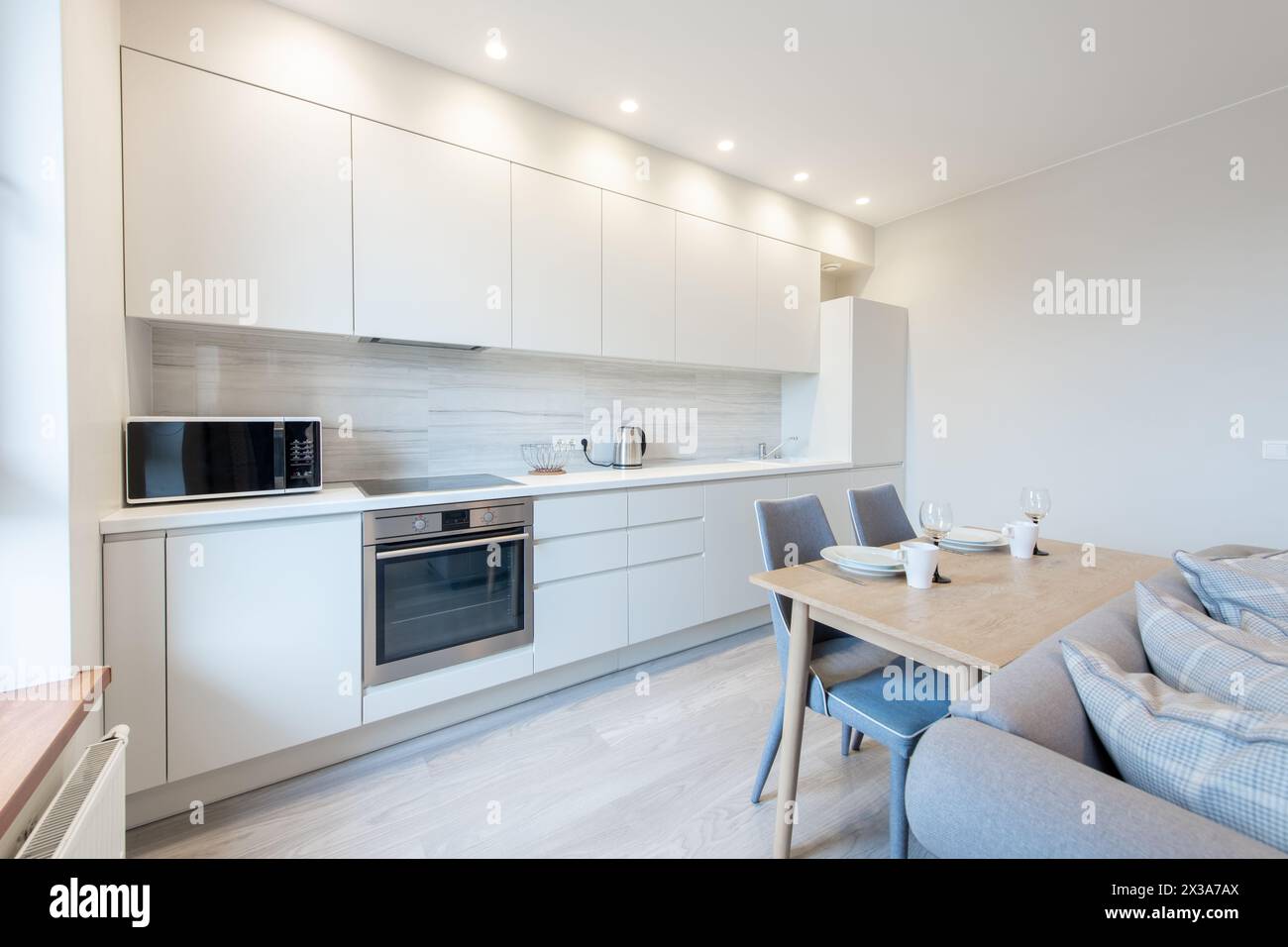 Classic White Kitchen Interior and Dinning Table. Oven, Kettle, Microwave and Glass. Plates on the Table. Minimalistic Interior Design. Stock Photo