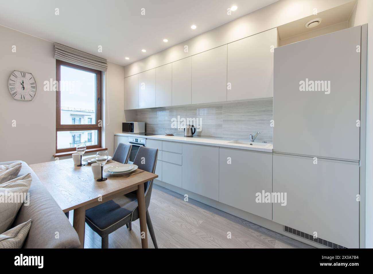 Classic White Kitchen Interior and Dinning Table. Oven, Kettle, Microwave and Glass. Plates on the Table. Minimalistic Interior Design. Stock Photo