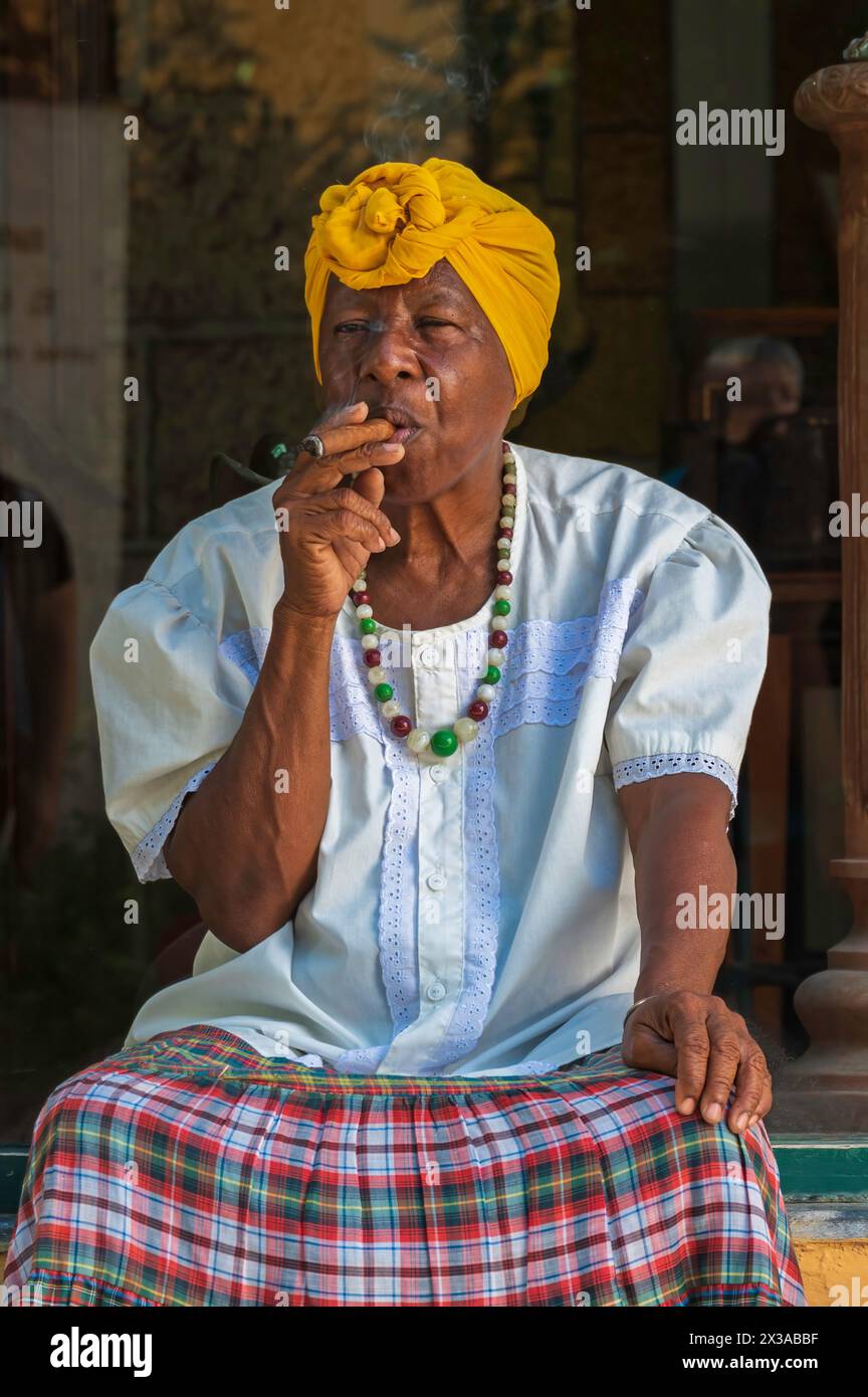 A lady sat on a window sill outside of a shop smoking a large cigar for tourists as a way of begging in the Old Town, Havana, Cuba. Stock Photo
