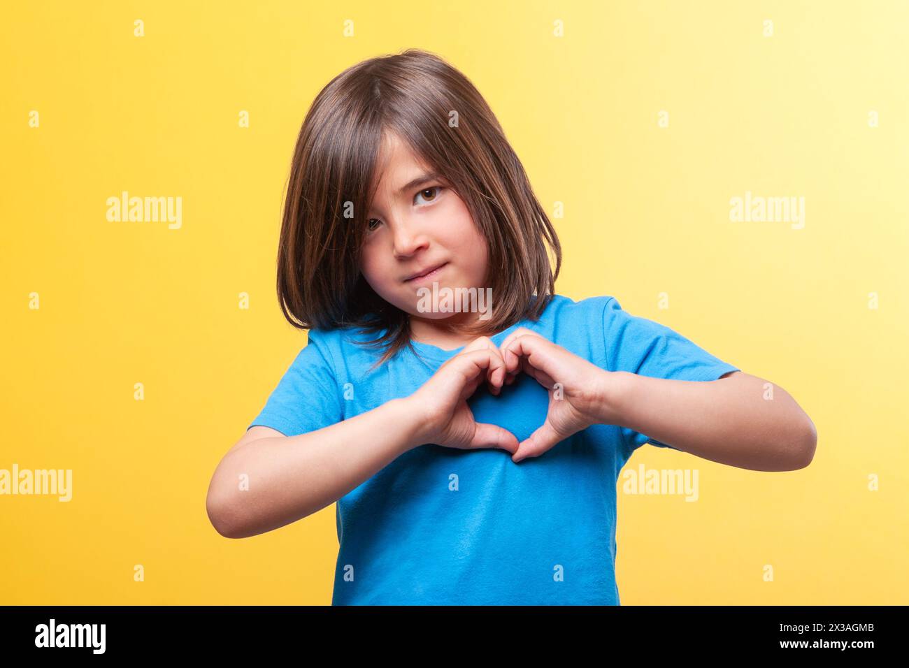 Boy with blue t-shirt and long brown hair makes the shape of a heart with his hands next to his chest while looking at the camera. Stock Photo