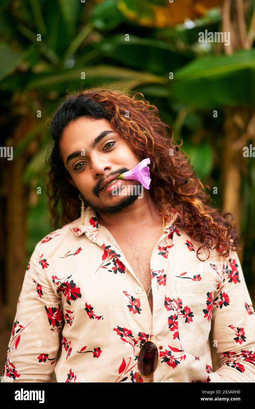 Gay man with curly hair and floral shirt stands in garden, purple flower in mouth. Expresses freedom, LGBT pride, gender fluidity. Relaxed, confident Stock Photo