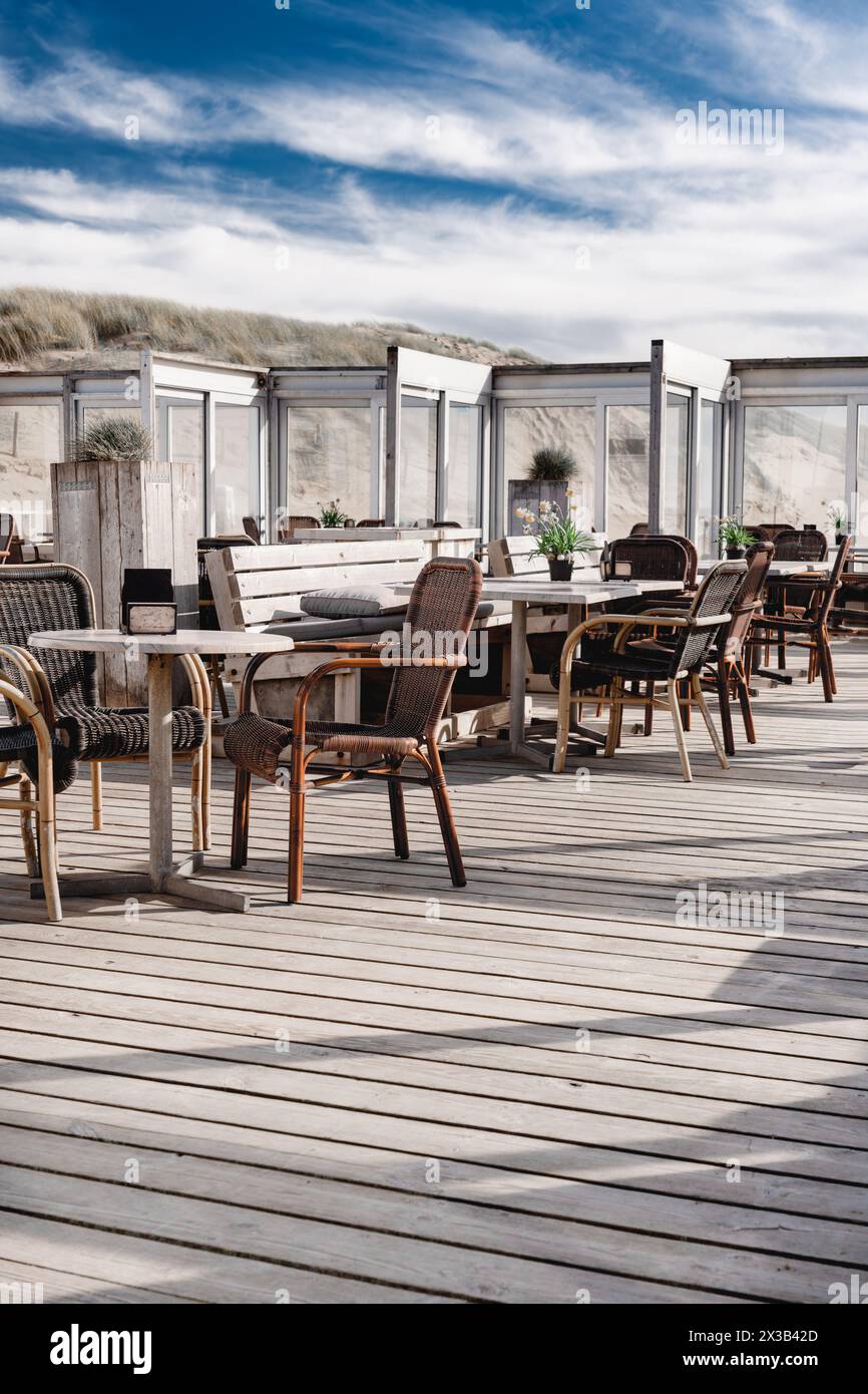 Beachside pavilion terrace overlooking the sand dunes on a soft blue cloud sky day. Vacant terrace of a beach pavilion, with empty chairs and tables Stock Photo