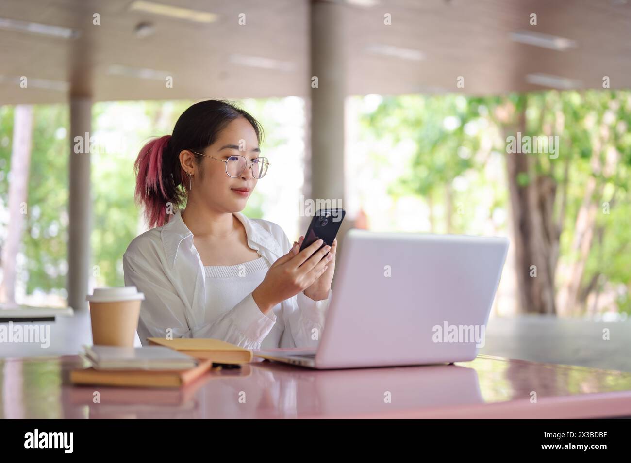 A young positive Asian female college student using her smartphone to check messages while sitting at a table in a campus cafeteria or canteen. Stock Photo