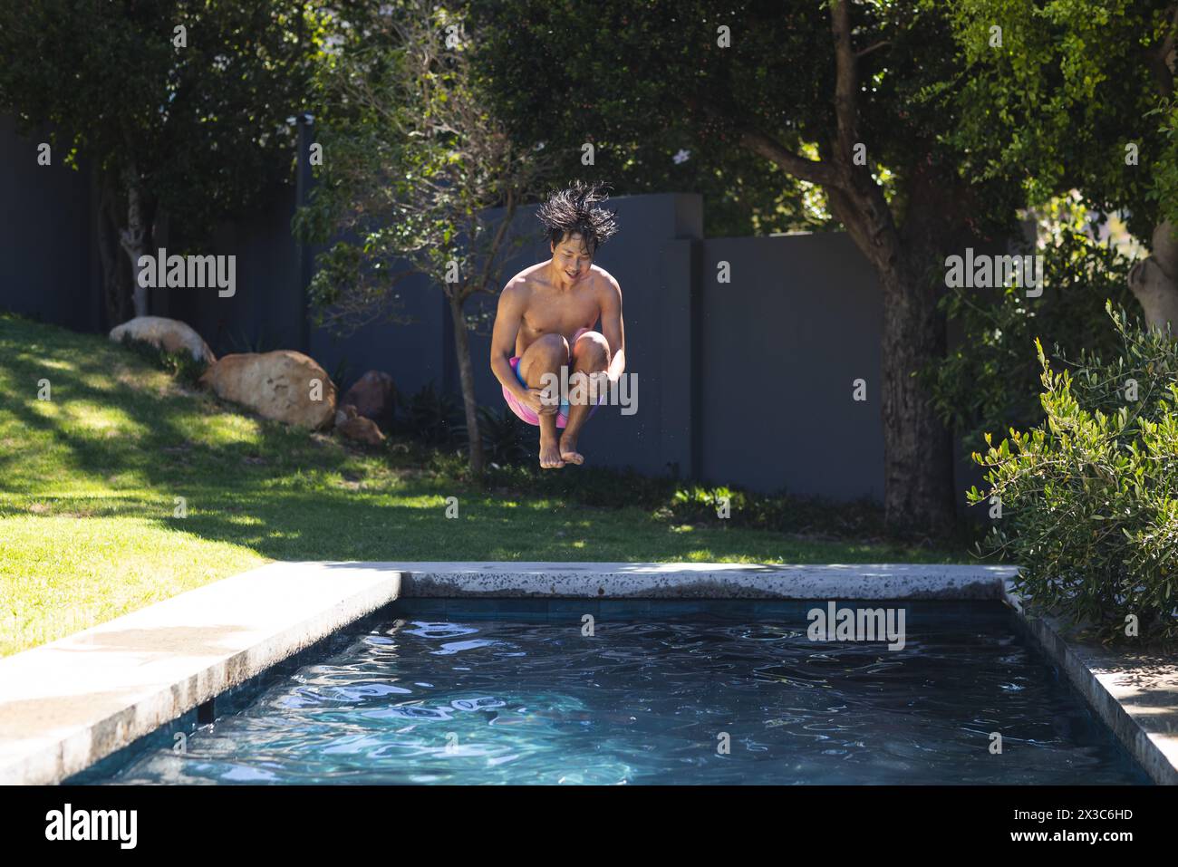 A teenage Asian boy is jumping into a pool at his home, wearing purple swim shorts, with copy space Stock Photo