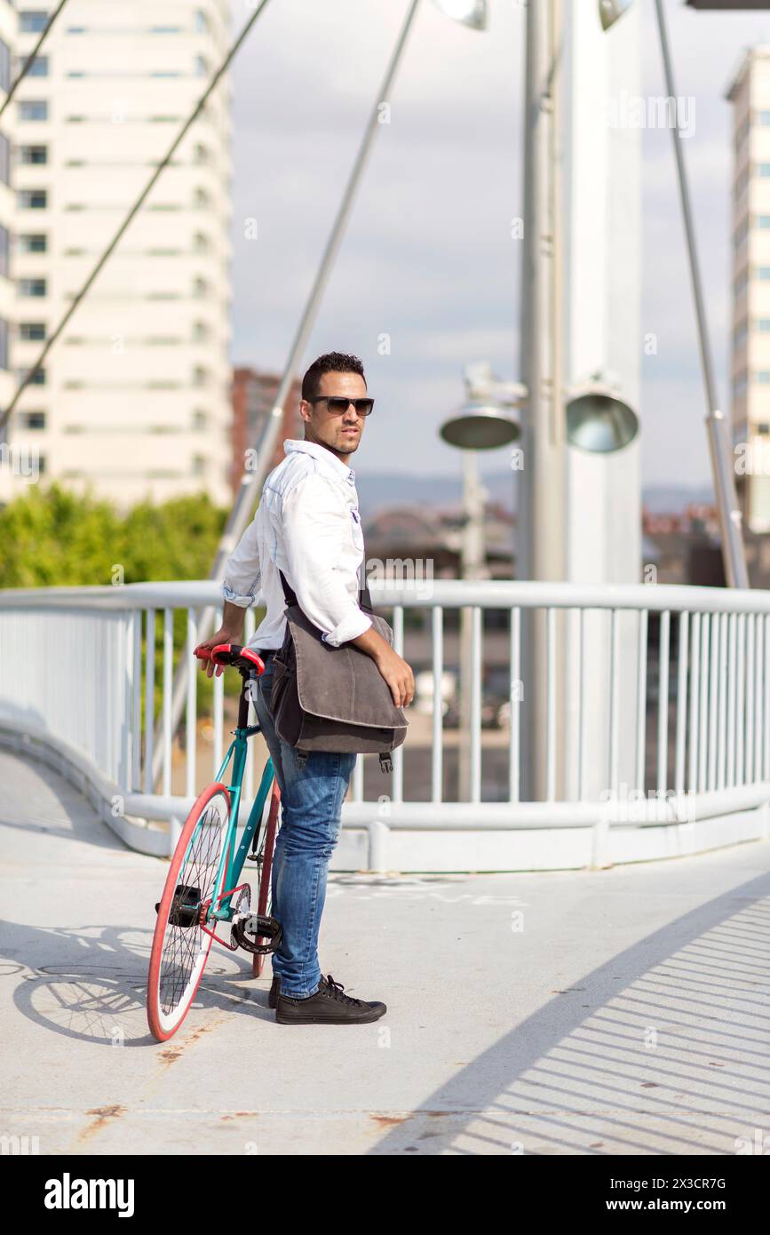 Casual man in sunglasses stands with his fixed-gear bike on a modern bridge, cityscape behind him. Stock Photo