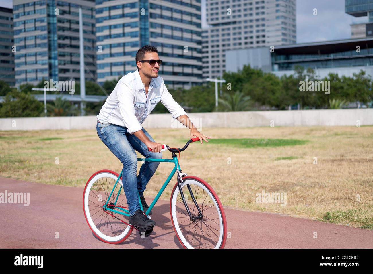 Fashionable young man riding a fixie bike in a city park, with modern buildings in the background, exemplifying eco-friendly commuting Stock Photo