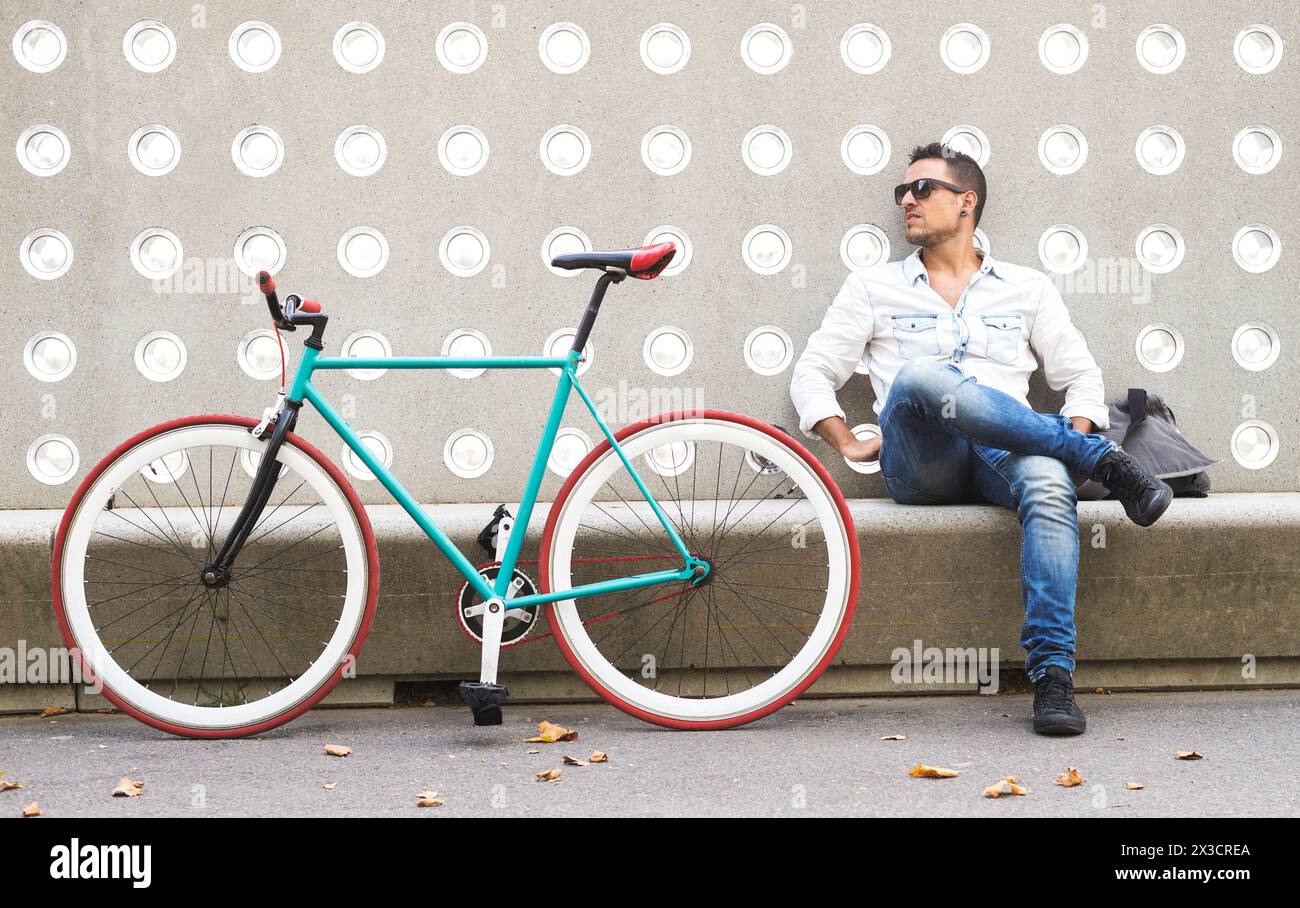 A trendy man in casual attire takes a break beside his colorful fixed-gear bike against a decorative urban wall Stock Photo