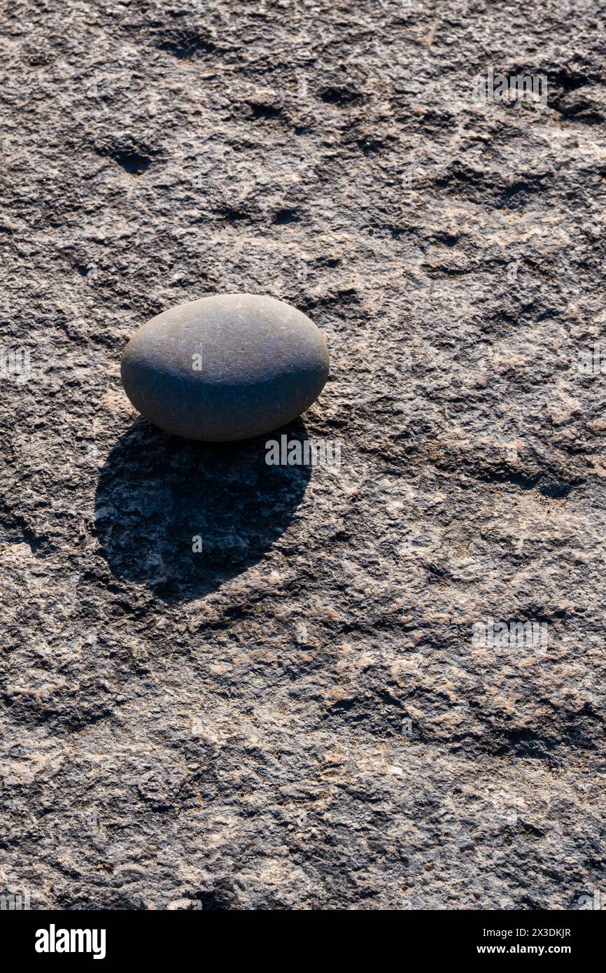 A smooth rounded pebble lying on the rough surface of a rock in Cornwall in the UK. Stock Photo