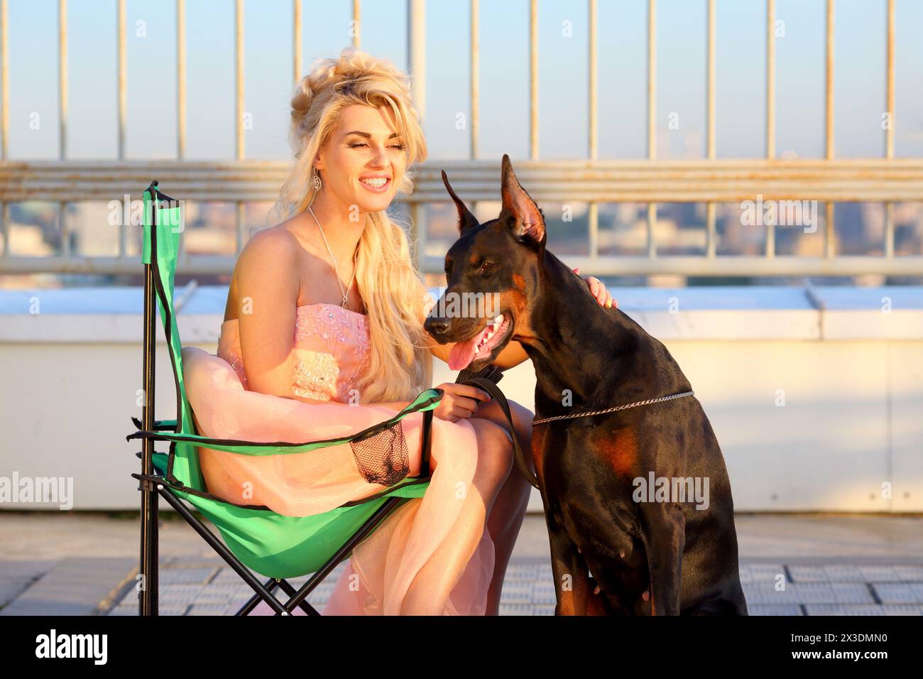 Happy beautiful blonde in a pink dress sitting on a folding chair near the Doberman on the roof Stock Photo