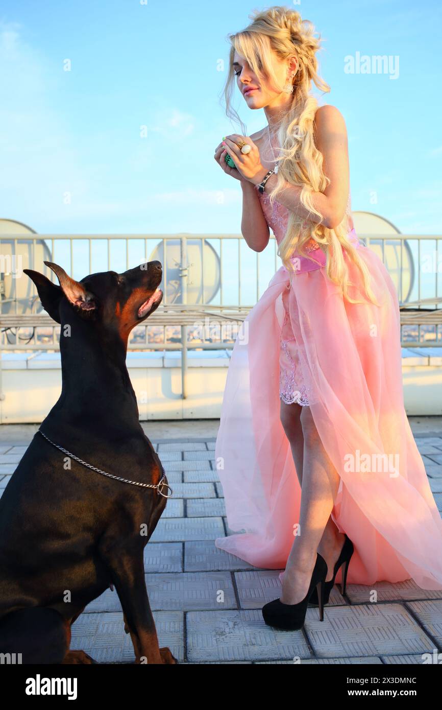 Beautiful girl with long blond hair in a pink dress playing with the Doberman on the roof Stock Photo