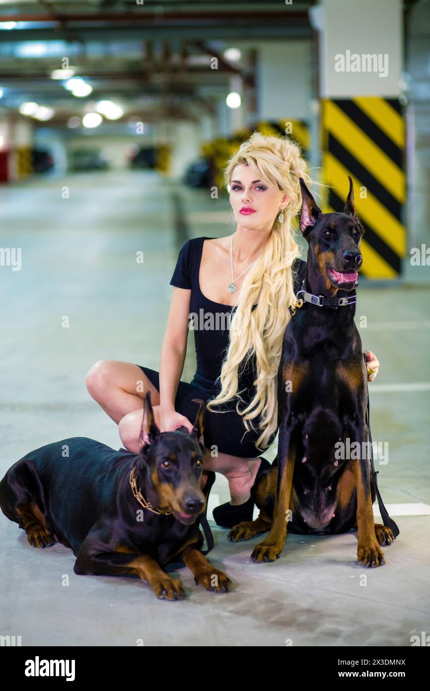Blonde in a black dress crouched in an embrace with two Doberman on leashes in the underground parking Stock Photo