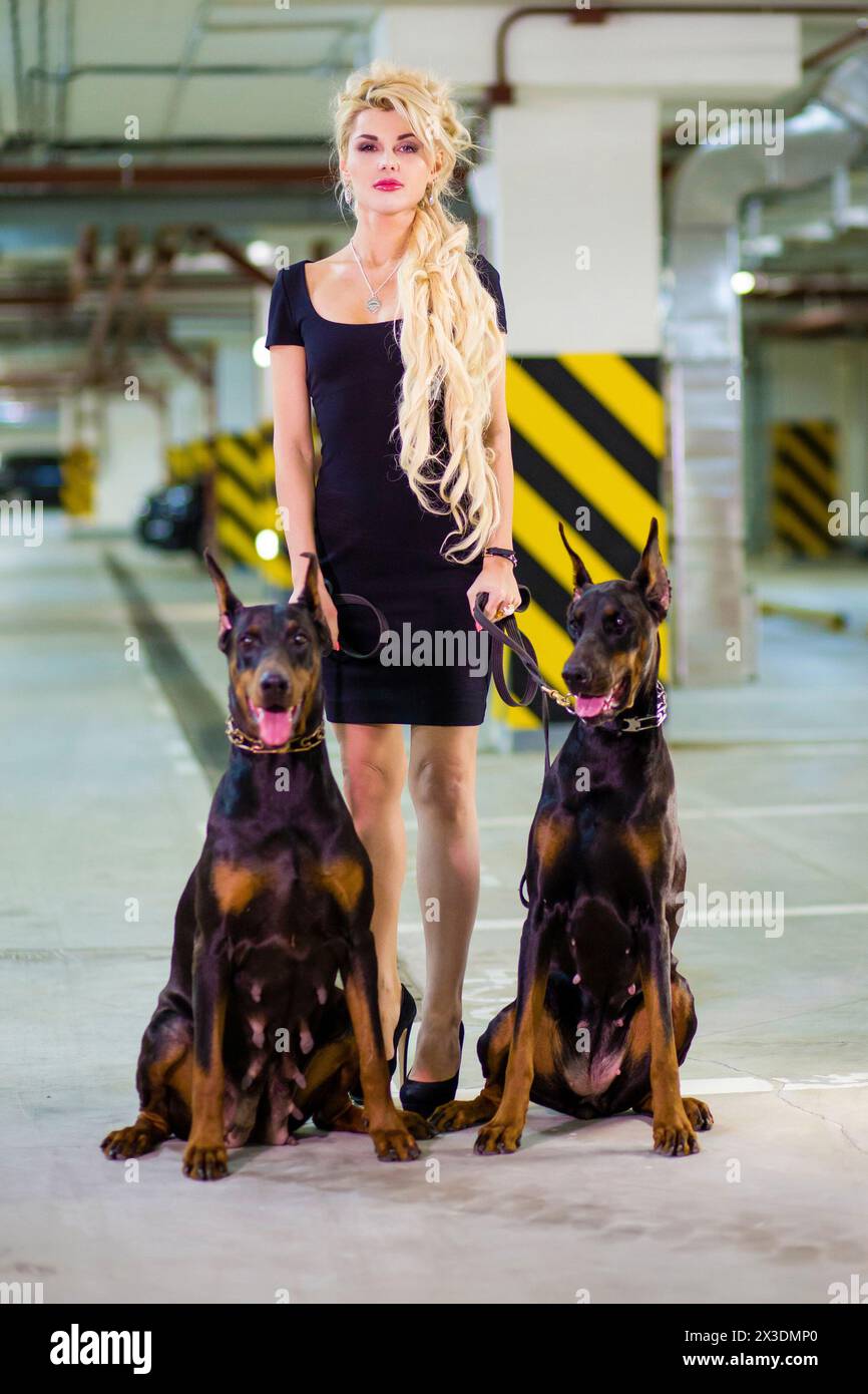 Portrait of beautiful woman in a black dress with two Doberman on leashes in the underground parking Stock Photo