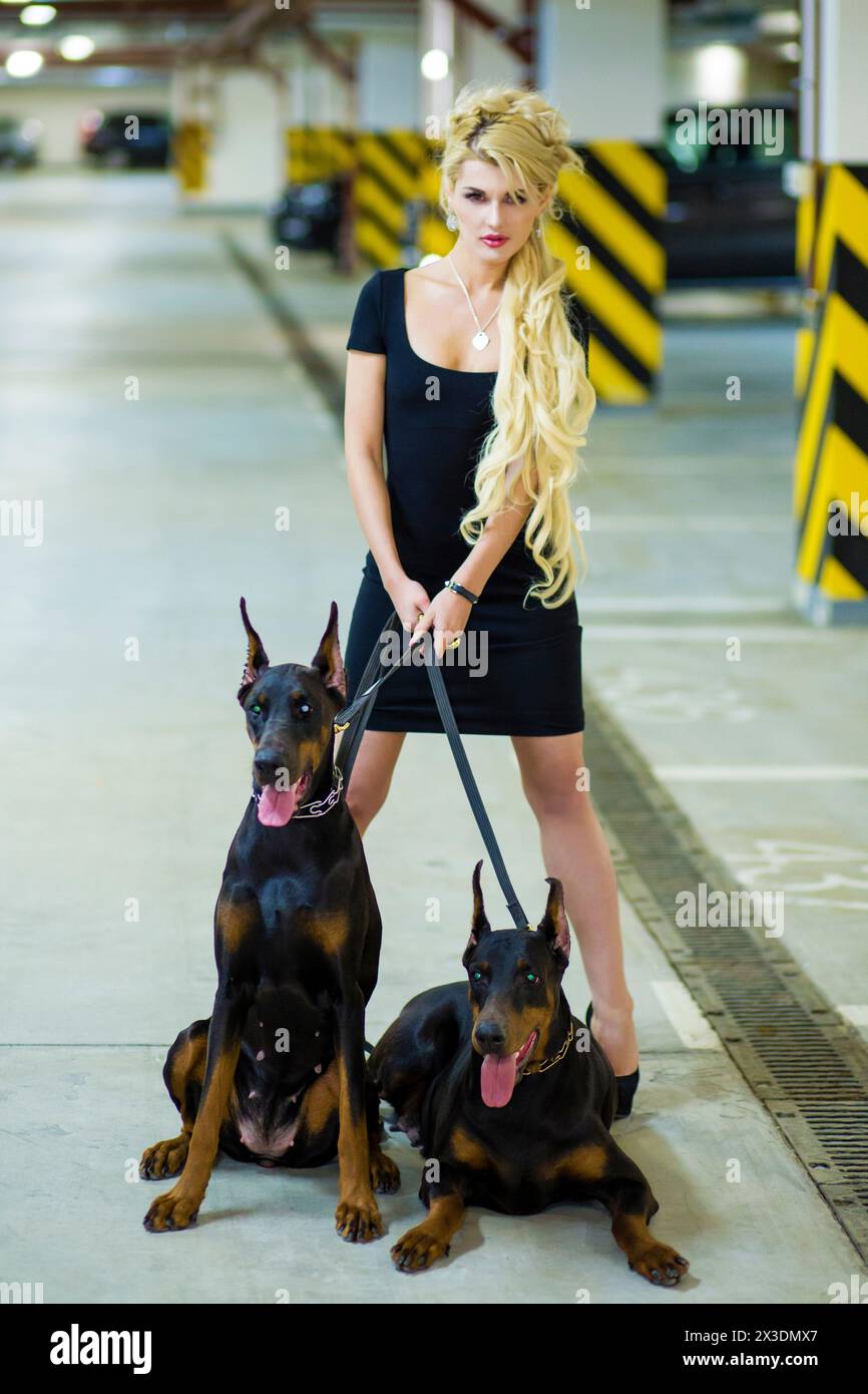 Beautiful woman with blond hair standing with two Doberman on leashes in the underground parking. Focus on dog Stock Photo