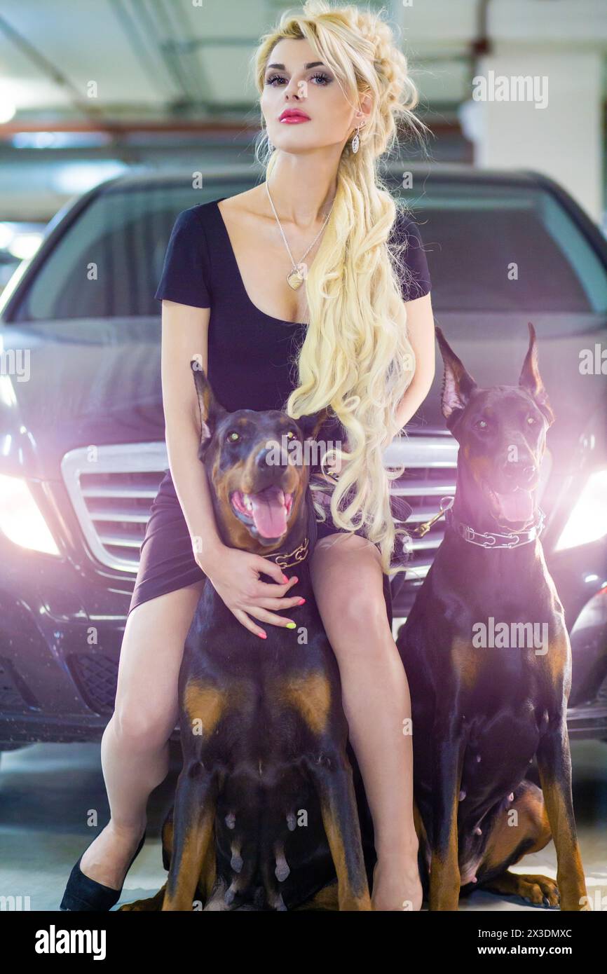 MOSCOW - JUN 01, 2015: Portrait of beautiful woman (with model release) with long blond hair in a black dress sitting on dog near black car with light Stock Photo