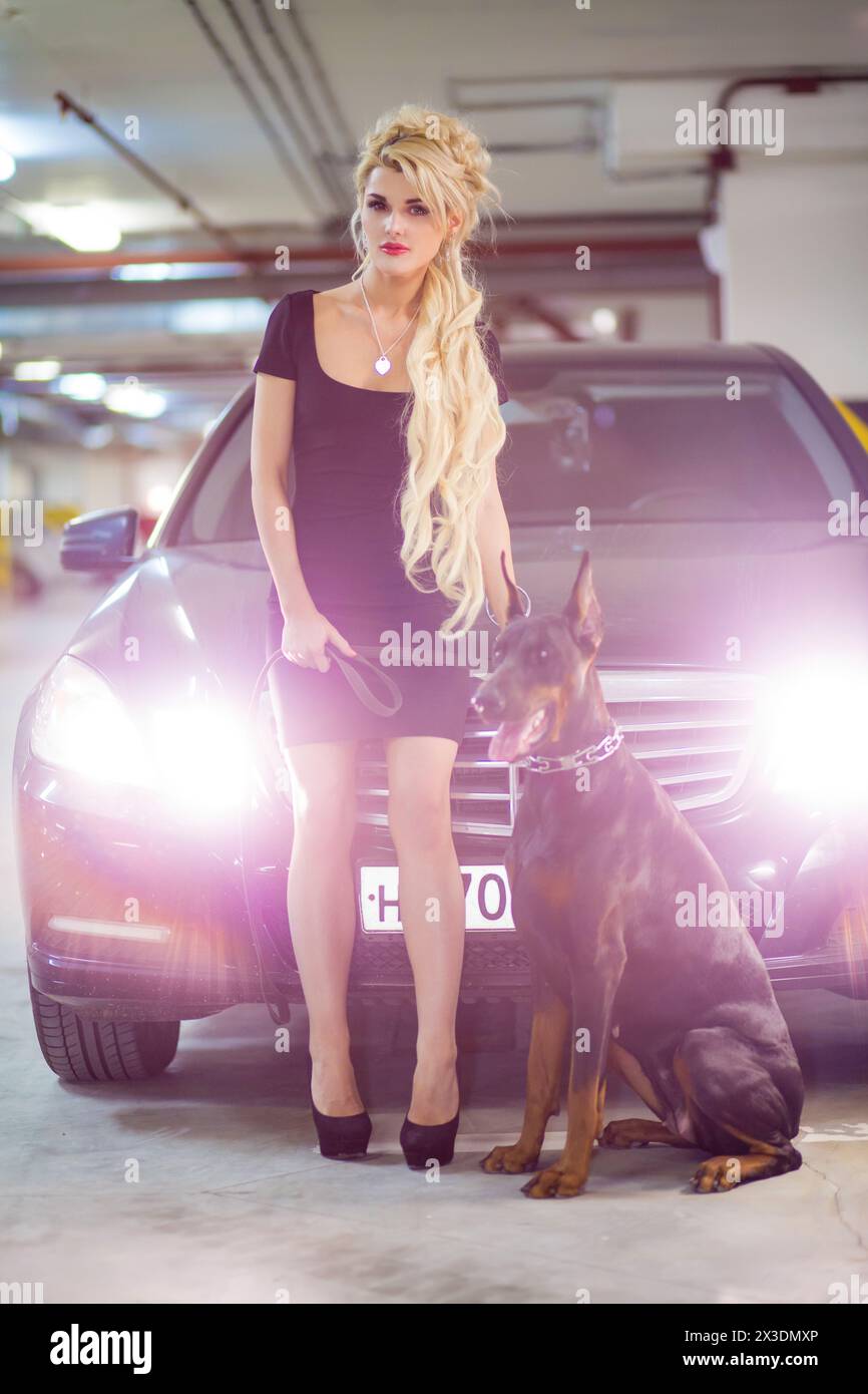 MOSCOW - JUN 01, 2015: Beautiful girl (with model release) with long blond hair in a black dress with dog standing near black car with lights on Stock Photo