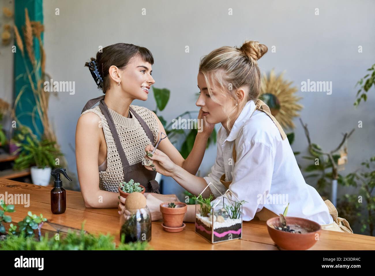 Two arty women sit at a table surrounded by potted plants, creating together with loving tenderness. Stock Photo